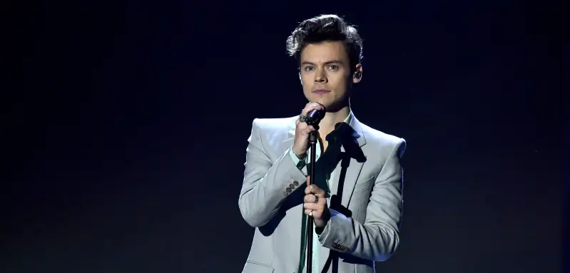Harry Styles steht auf der Bühne mit Mikrofon in der Hand vor einem dunklen Hintergrund