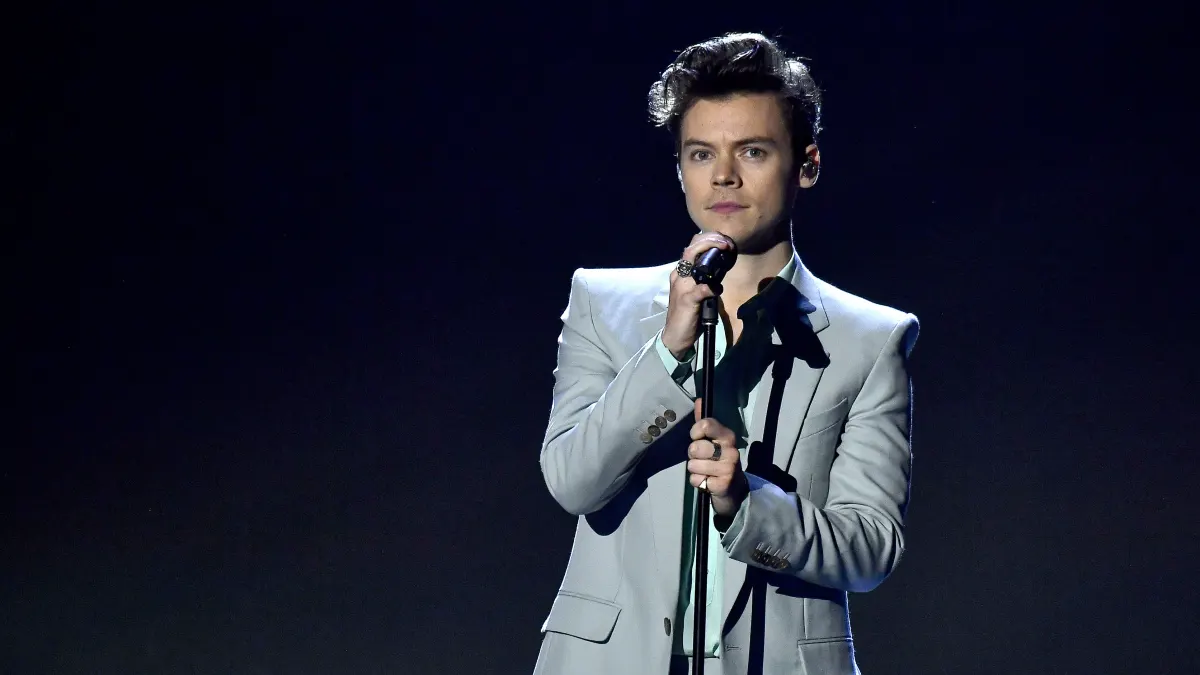 Harry Styles steht auf der Bühne mit Mikrofon in der Hand vor einem dunklen Hintergrund