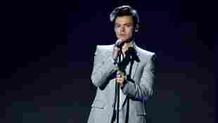Harry Styles steht auf der Bühne mit Mikrofon in der Hand vor einem dunklen Hintergrund