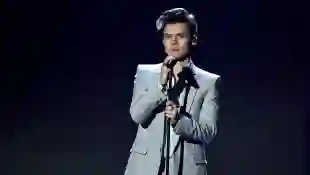 Harry Styles steht auf der Bühne mit Mikrofon in der Hand vor einem dunklen Hintergrund