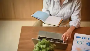 Cropped shot of female manager holding her personal daily planner and using laptop at desk. Model Re