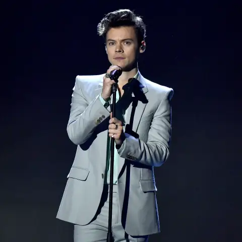Harry Styles steht auf der Bühne mit Mikrofon in der Hand vor einem dunklen Hintergrund