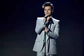 Harry Styles steht auf der Bühne mit Mikrofon in der Hand vor einem dunklen Hintergrund