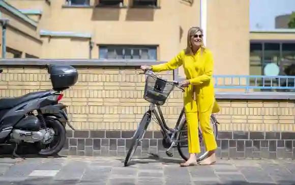 Königin Máxima der Niederlande strahlt in gelbem Outfit neben ihrem Fahrrad auf dem Weg zu einem Ausstellungsbesuch.