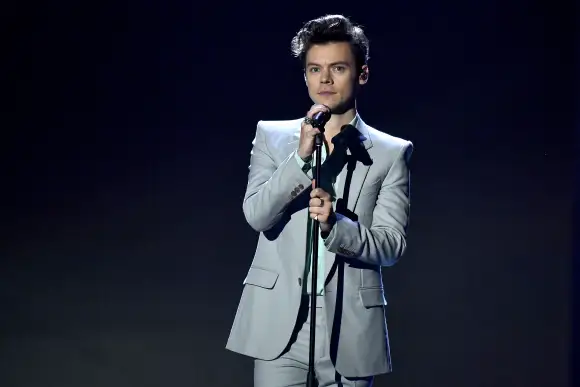 Harry Styles steht auf der Bühne mit Mikrofon in der Hand vor einem dunklen Hintergrund